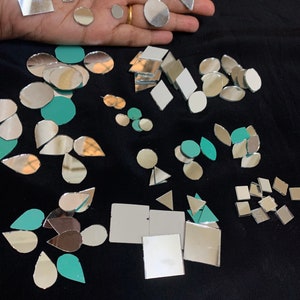 May include: Assortment of small mirror tiles in various shapes and sizes, including circles, ovals, squares, diamonds, and teardrops. The tiles are arranged on a black surface. Some of the tiles are silver, while others are a light teal color.