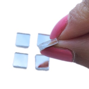 May include: Four small square mirror tiles, one of which is being held by a finger with pink nail polish.