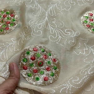 Puede incluir: Tela beige con diseños florales bordados en blanco y adornos de lentejuelas doradas, verdes y rojas en forma de círculos.