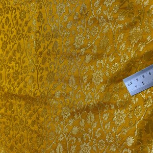 May include: A close-up of a yellow fabric with a gold floral pattern. A ruler is placed on the fabric to show the scale.