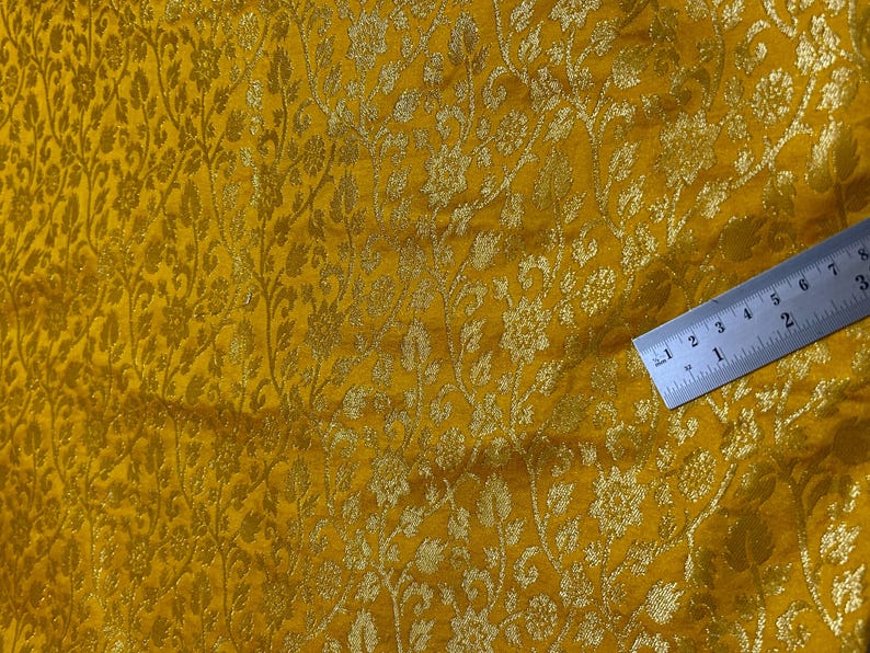 May include: A close-up of a yellow fabric with a gold floral pattern. A ruler is placed on the fabric to show the scale.