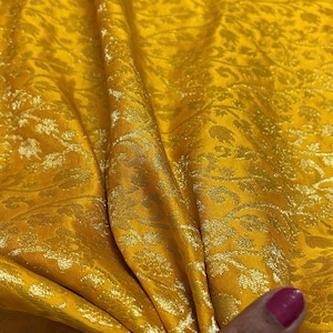 May include: A yellow fabric with a gold floral pattern. The fabric is folded and draped over a hand.