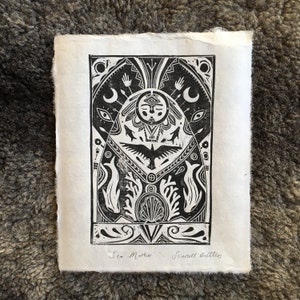 May include: Black and white woodcut print of a stylised sea creature with a human face, surrounded by geometric patterns and symbols. The print is titled "Sea Mother" and signed "Scarlett Butters".