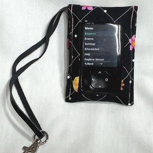 May include: A black fabric pouch with a floral pattern and a clear plastic window containing a digital device. The screen displays text including Menu, Reports, Events, Settings, Information, Help, and Replace Sensor. A black strap with a metal clip is attached.
