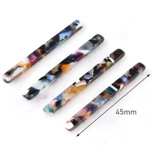 Könnte beinhalten: Vier farbige, rechteckige Haarklammern, jede mit einem anderen Muster aus Schwarz, Weiß, Blau, Orange und Gelb. Die Klammern sind 4,5 cm lang.