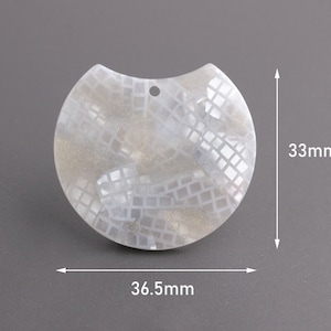 Może przedstawiać: Biały, półokrągły wisiorek z geometrycznym wzorem i otworem na górze. Wisiorek ma 36,5 mm szerokości i 33 mm wysokości.