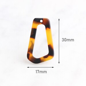 Könnte beinhalten: Ein Anhänger aus Acryl mit Schildpattmuster und einer einzigartigen, offenen geometrischen Form. Der Anhänger ist 30 mm lang und 17 mm breit und hat ein Loch zum Auffädeln. Das Farbmuster umfasst Brauntöne und Gelb.