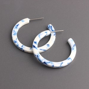 May include: A pair of white and blue marbled hoop earrings. The earrings are made of a thick, rounded plastic and have a silver post.