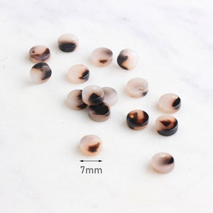 Puede incluir: Un conjunto de cuentas redondas, cada una de aproximadamente 7 mm de diámetro, con un patrón veteado en rosa claro y marrón oscuro. Las cuentas están dispersas sobre una superficie blanca, mostrando sus variaciones de color y diseño.