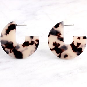 Puede incluir: Un par de pendientes de aro de carey blanco y negro. Los pendientes son de forma semicircular con un hueco en el medio.