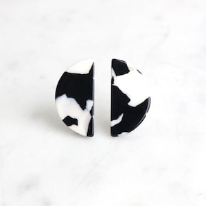 May include: A pair of half-circle stud earrings. Each earring features a black and white marbled pattern. The earrings are made of a glossy material and are set against a white background. The earrings are a modern accessory.