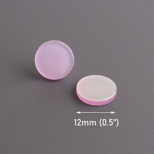May include: Two round, flat, pink and white cabochons, each approximately 0.5 inches (12mm) in diameter, rest on a gray surface. One cabochon is upright, while the other lies on its side, revealing a white interior.
