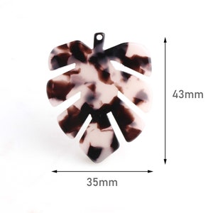 Puede incluir: Un dije de hoja de plástico moteado marrón y blanco con un agujero en la parte superior para colgar. La hoja mide 4,3 cm de alto y 3,5 cm de ancho.