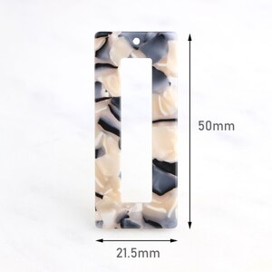 Puede incluir: Un colgante rectangular de plástico marmoleado blanco y negro con un agujero en la parte superior y un recorte rectangular en el medio. El colgante mide 21,5 mm de ancho y 50 mm de alto.