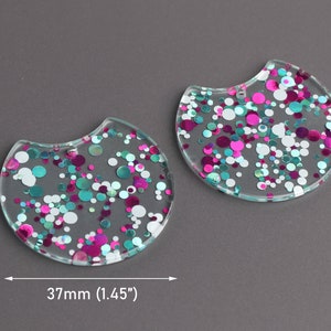 May include: Two clear, round, resin shapes with a scalloped edge. The shapes are filled with pink, teal, and white glitter. The shapes measure 1.45 inches in diameter.