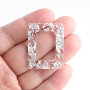 May include: A clear, rectangular resin pendant with a silver foil inlay. The pendant has a rectangular cutout in the center. The pendant is held in a person's hand against a white background.