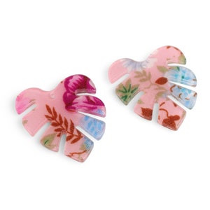 May include: Two pink, leaf-shaped earrings with a floral pattern. The earrings have a monstera leaf design with cut-out sections. The floral pattern includes shades of pink, purple, blue, and green. The earrings are set against a white background.