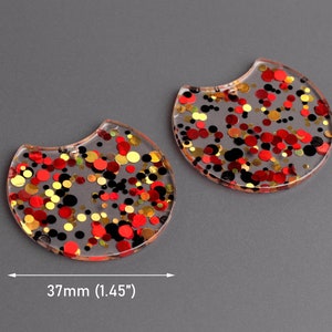 May include: Two clear resin earrings with a crescent shape. The earrings are filled with red, gold, and black glitter.