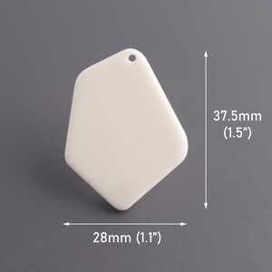 May include: A white, irregular-shaped pendant with a small hole at the top for stringing. The pendant measures 1.1 inches wide and 1.5 inches long. The background is a solid gray.