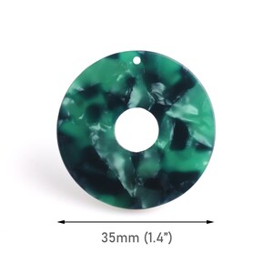Puede incluir: Un colgante en forma de rosquilla de acrílico veteado verde y negro, con un agujero en el centro. El colgante mide 3,5 cm de diámetro y tiene un pequeño agujero en la parte superior para ensartar. La superficie es brillante.