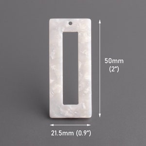 Könnte beinhalten: Ein rechteckiger, perlmuttfarbener weißer Acryl-Anhänger mit hohler Mitte. Der Anhänger ist 50 mm lang und 21,5 mm breit, mit einem kleinen Loch oben zum Auffädeln. Das Material hat einen marmorierten Effekt.