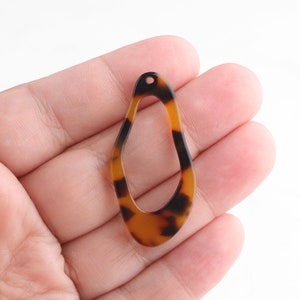 May include: A teardrop-shaped earring component with a tortoiseshell pattern. The open-center pendant is brown and black with a small hole at the top for attachment. The earring component is held in a hand.