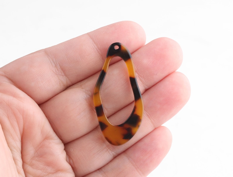 May include: A teardrop-shaped earring component with a tortoiseshell pattern. The open-center pendant is brown and black with a small hole at the top for attachment. The earring component is held in a hand.