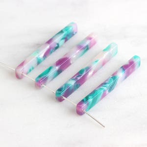 May include: Four long, thin, rectangular acrylic beads with a marbled pattern in shades of white, pink, and teal. The beads are strung on silver wires.
