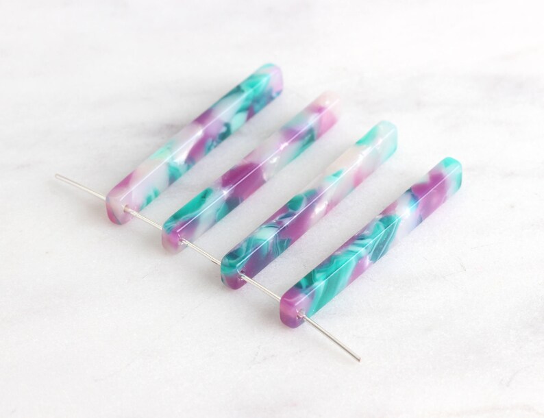 May include: Four long, thin, rectangular acrylic beads with a marbled pattern in shades of white, pink, and teal. The beads are strung on silver wires.