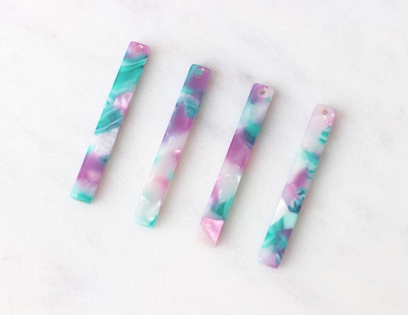 May include: Four colorful, rectangular, acrylic charms with a swirl pattern in shades of pink, green, and white. Each charm has a hole at the top for attaching to jewelry or other crafts.