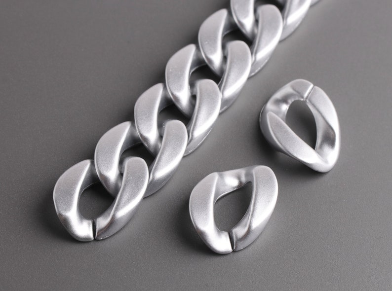 Pu&ograve; includere: Una collana a catena argentata con grandi maglie spesse. La catena &egrave; realizzata in metallo liscio e lucido e presenta un design moderno e minimalista.