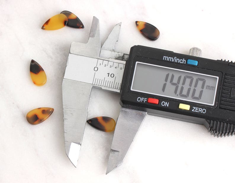 May include: A close-up of a digital caliper measuring a small, brown and tan, teardrop-shaped object. The caliper reads 14.00 mm.