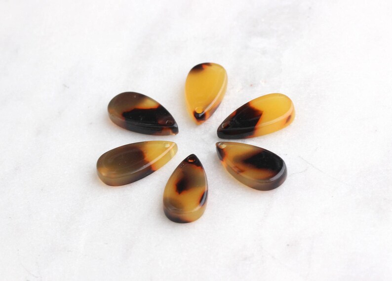 May include: Six brown and yellow teardrop-shaped pendants with a hole at the top. The pendants are arranged in a circle on a white surface.