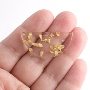 Puede incluir: Un colgante de resina transparente en forma de semic&iacute;rculo con escamas de pan de oro incrustadas en el interior.