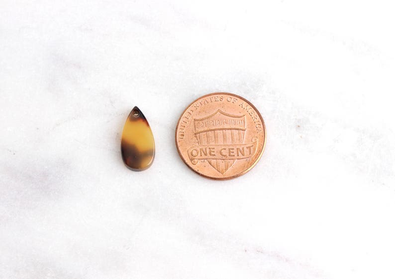 May include: A small, teardrop-shaped pendant made of brown and tan tortoiseshell material. The pendant is about the size of a US penny.