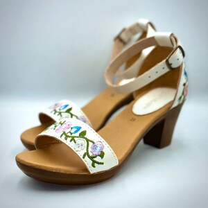 Puede incluir: Sandalias de cuero blanco con un dise&ntilde;o de bordado floral. Las sandalias tienen un tac&oacute;n de madera marr&oacute;n y una correa de tobillo con cierre de hebilla.