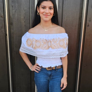 Puede incluir: Una mujer con un top blanco de hombros descubiertos con bordados dorados y jeans azules. Est&aacute; de pie frente a una puerta de madera.