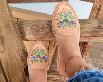 Puede incluir: Un par de sandalias de cuero color canela con flores bordadas de colores en la punta. Las sandalias tienen correas para el tobillo y las lleva una persona que lleva pantalones vaqueros azules.