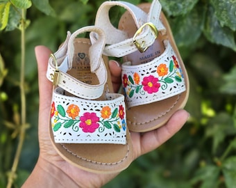 Può includere: Un paio di sandali da bambino in pelle bianca con ricami floreali colorati. I sandali hanno una chiusura con fibbia e una suola marrone. I sandali sono tenuti in mano su uno sfondo di fogliame verde.