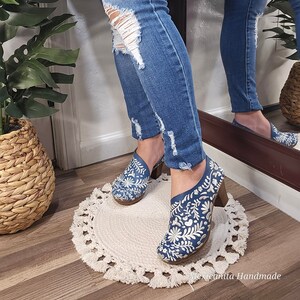 May include: Blue denim shoes with a floral embroidery design and a wooden heel. The shoes are paired with distressed blue jeans. The shoes are on a small, round, cream-colored rug with a tassel trim. The image is well-lit and shows the shoes in detail.