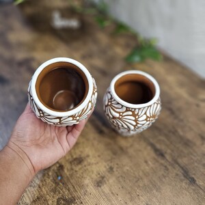 May include: Two small, round, brown ceramic pots with white floral designs. The pots have a wide opening and are held in a person's hand. The pots are on a wooden surface. The text "Mexicanita Handmade" is visible.