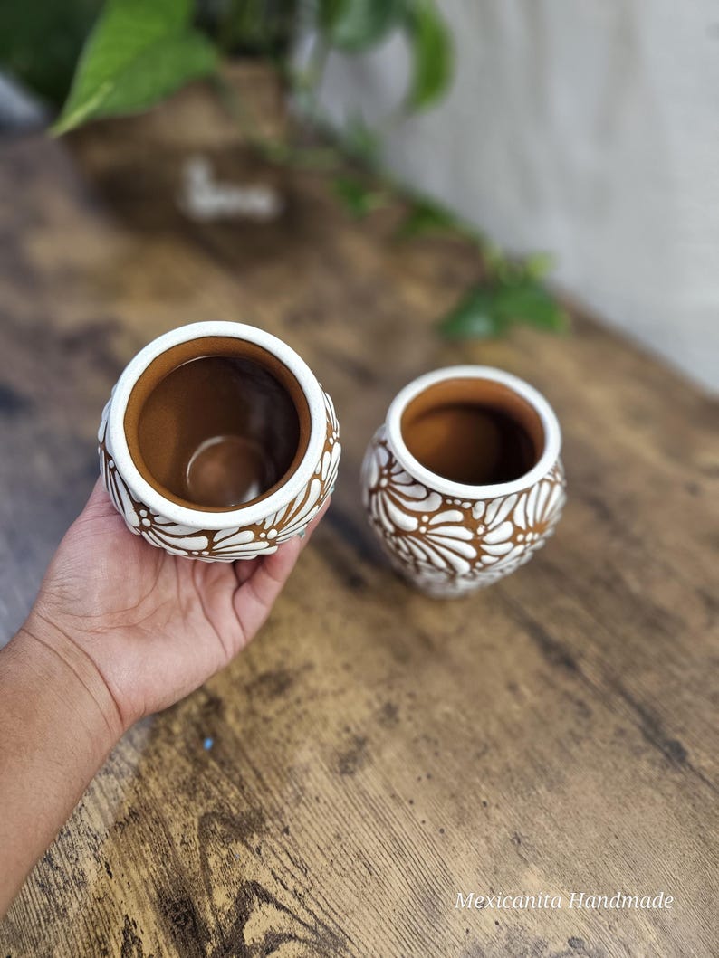 May include: Two small, round, brown ceramic pots with white floral designs. The pots have a wide opening and are held in a person's hand. The pots are on a wooden surface. The text "Mexicanita Handmade" is visible.