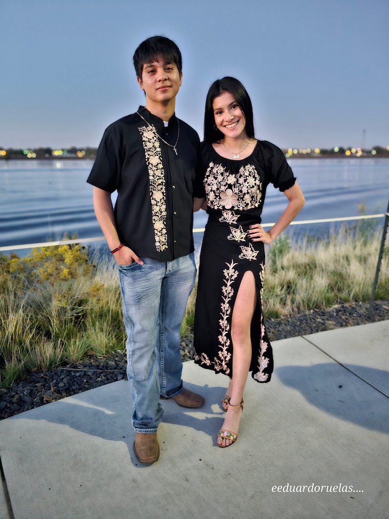 May include: A couple poses for a photo in front of a body of water. The woman is wearing a black dress with gold embroidery and a slit up the side. The man is wearing a black button-down shirt with gold embroidery and blue jeans.