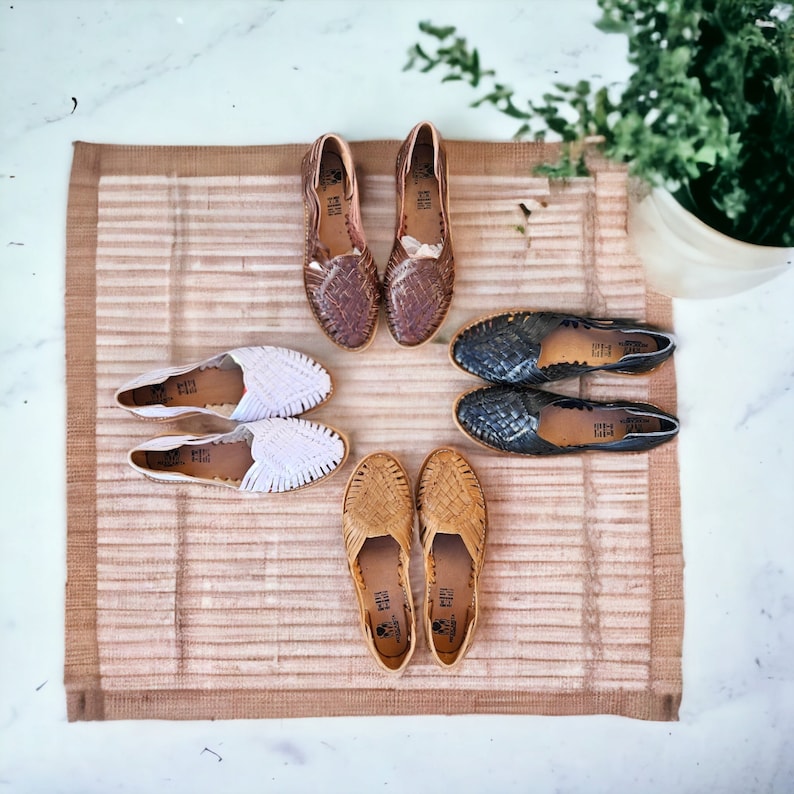 Puede incluir: Un conjunto de ocho sandalias de cuero tejido en varios colores, incluyendo marr&oacute;n, negro, blanco y beige. Las sandalias est&aacute;n dispuestas sobre una estera tejida con un patr&oacute;n de rayas marrones y blancas.
