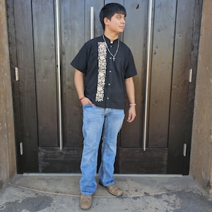 May include: A young person wearing a black short-sleeved shirt with a white embroidered design and blue jeans. The shirt has a button-up collar and a silver chain necklace. The person is standing in front of a wooden door.