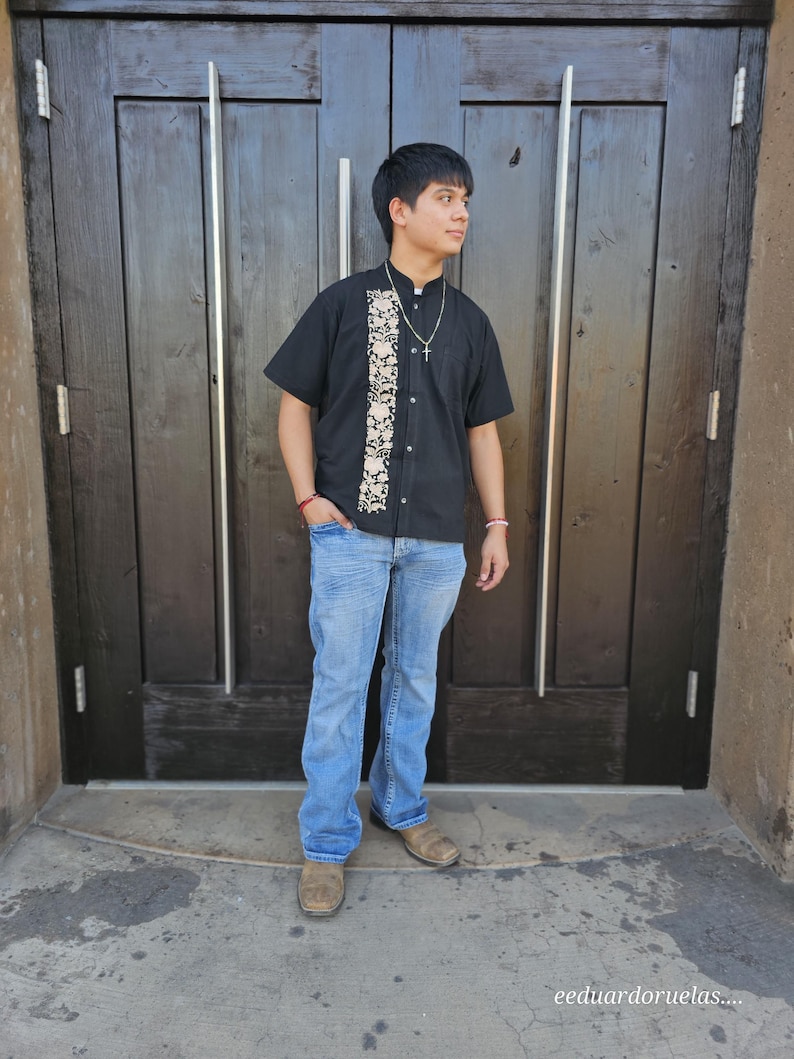 May include: A young person wearing a black short-sleeved shirt with a white embroidered design and blue jeans. The shirt has a button-up collar and a silver chain necklace. The person is standing in front of a wooden door.