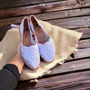 Puede incluir: Un par de sandalias de cuero blanco con tiras tejidas. Las sandalias tienen una punta puntiaguda y un tac&oacute;n bajo. Las sandalias est&aacute;n sobre una superficie marr&oacute;n claro.