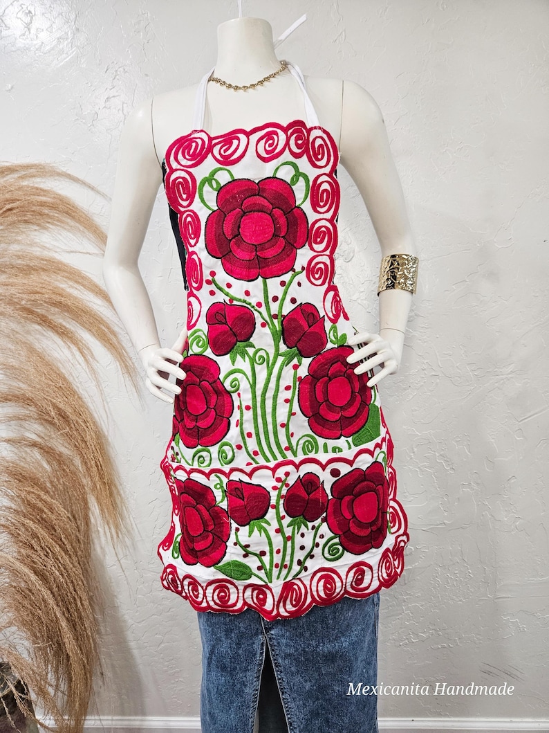 Puede incluir: Delantal blanco con un dise&ntilde;o de bordado floral rojo y verde. El delantal tiene un borde festoneado y una correa de cuello halter.