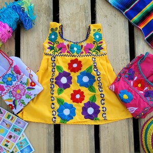 Pode incluir: Um vestido amarelo sem mangas com bordado floral colorido. O vestido tem um decote redondo e uma saia franzida. O vestido est&aacute; sobre uma superf&iacute;cie de madeira com outros itens, incluindo uma pi&ntilde;ata, um pequeno vestido branco e um vestido rosa.