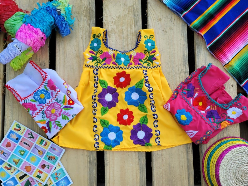 Pode incluir: Um vestido amarelo sem mangas com bordado floral colorido. O vestido tem um decote redondo e uma saia franzida. O vestido est&aacute; sobre uma superf&iacute;cie de madeira com outros itens, incluindo uma pi&ntilde;ata, um pequeno vestido branco e um vestido rosa.